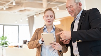 Zwei Personen stehen nebeneinander und blicken auf ein Tablet, im Hintergrund ist ein modernes und hell beleuchtetes Büro