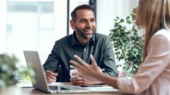 Person mittleren Alters mit grünem Hemd sitzt an einem Tisch mit einem Laptop und unterhält sich mit einer anderen Person