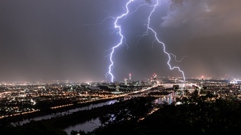 Panoramaaufnahme einer Stadt bei Nacht. Die Straßen sind beleuchtet. Der Himmel ist bewölkt. Am Himmel sind mehrere, große, leuchtende Blitze