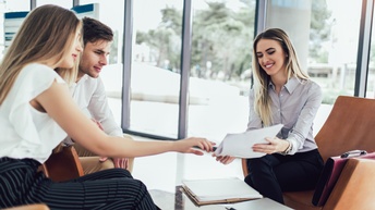Um einen niedrigen Tisch sitzen drei Personen. Die Person rechts hält den beiden Personen links einen Zettel hin, auf den alle drei blicken. Im Hintergrund ist eine Glasfront