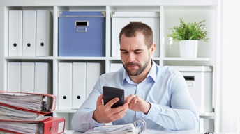 Frontale Aufnahme einer Person in einem hellblauen Hemd, die an einem Schreibtisch sitzt und auf einem Taschenrechner tippt. Vor ihr liegt ein aufgeklappter Ordner mit Unterlagen. Daneben sind weitere, geschlossene, dicke Ordner