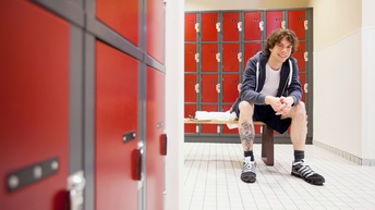 Lächelnde Person sitzt in Umkleidebereich umgeben von roten Spinden