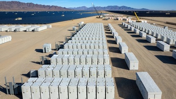 Vogelperspektive auf ein Feld voller Batteriespeicher, im Hintergrund breitflächige Solaranlagen und Bergkette unter blauem Himmel
