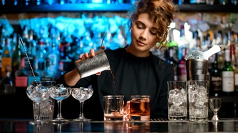 Person steht hinter einer Bar und schenkt einen Cocktail ein, während auf der Theke davor verschiedene Gläser gefüllt mit Eis stehen 