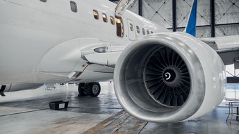 Nahaufnahme der Turbine eines Flugzeugs. In der Turbine ist ein sich eindrehendes Gitter. Das Flugzeug ist weiß. Eine Tür auf der Seite ist offen. Das Flugzeug steht in einer Halle auf einem asphaltierten Boden