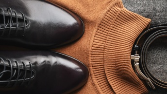 Topshot eines hellbraunen Pullovers, auf dem ein Paar schwarzer Lederschuhe sind. Neben dem Pullover liegt ein eingedrehter Gürtel