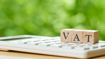 Detailansicht eines Taschenrechners auf dem Holzwürfel mit schwarzen Buchstaben liegen, die den Schriftzug VAT ergeben, Hintergrund verschwommen grün