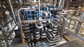 In einer Produktionsstätte stehen verschiedene große Maschinen mit Rohren und Verbindungen.