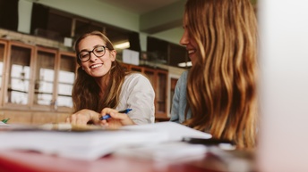 Zwei lächelnde junge Personen sitzen in Lesesaal und blicken auf Unterlagen vor sich