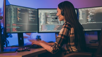 Lächelnde Person mit Headset sitzt vor mehreren Monitoren und programmiert