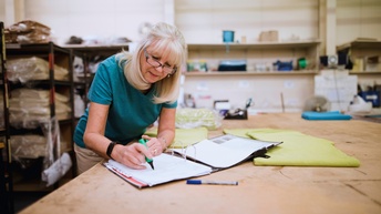 Person mit blonden Haaren und Brille mittleren Alters steht in einer Schneiderei und markiert Blätter einer geöffneten Arbeitsmappe während ein grüner Stoff neben ihr auf der Arbeitsfläche liegt. 