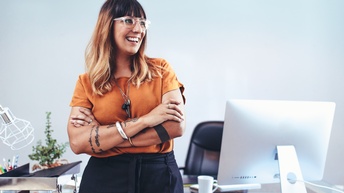 Frontale Aufnahme einer Person in einem orangen Oberteil, dunkler Hose, verschränkten Armen und Brille, die in den rechten Bildbereich blickt. Die Person lehnt sich an einem Schreibtisch hinter ihr an, auf dem ein Bildschirm und Büroutensilien sind.