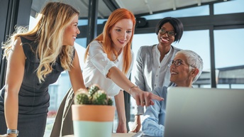 An einem Tisch sitzt eine Person mit Brille und kurzen weißen Haaren. Vor ihr steht ein Laptop. Neben und hinter ihr stehen drei Personen. Die mittlere Person deutet auf den Bildschirm des Laptops. Alle anderen blicken zu ihr