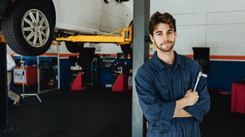 Person in blauem Overall mit Schraubenzieher in der Hand in Werkstatt stehend im Hintergrund Hebetribüne mit Auto