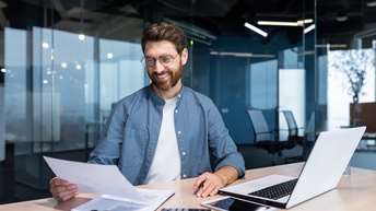Person mit dunklen kurzen Haaren, Bart und Brille sowie Hemd sitzt an einem Schreibtisch mit Laptop, Tablet, Taschenrechner und blickt auf ein Dokument, im Hintergrund spiegelnde Glasfronten, die das Büro eingrenzen