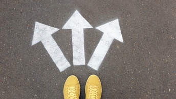 Gelbe Schuhe stehen auf einem Asphalt, davor sind drei weiße Pfeile, die in unterschiedliche Richtungen zeigen am Boden markiert