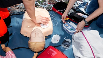 Nahaufnahme Puppe, die am Boden liegt. Die Hände einer Person liegen aufeinander gepresst auf dem Brustbereich der liegenden Puppe. Rechts davon sind die Hände einer weiteren Person, die einen Defibrillator umfassen.