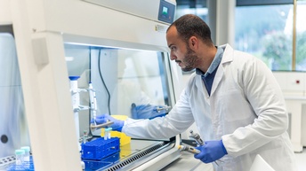 Seitliche Aufnahme einer Person mit weißem Kittel, Bart, dunklen Haaren und Handschuhen. Die Person steht an einer Station mit einer Glasscheibe, hinter der Reagenzgläser stehen