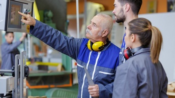 Zwei jüngere Personen in Arbeitskleidung blicken auf Display einer Maschine, auf das ältere Person in Arbeitskleidung mit Hörschutz um den Hals deutet