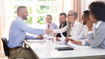 Seitliche Aufnahme eines Tisches. Links am Tisch sitzt eine Person. Ihr gegenüber sitzen fünf Personen. Die Person links und eine Person rechts schütteln sich die Hände und lächeln