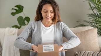 Frontalaufnahme einer auf einem Sofa sitzenden Person in hellem Oberteil und grauer Weste, die auf dem Schoß einen kleinen, geöffneten Karton hat, in dem ihre beiden Hände verschwinden.  Die Person blickt auf den geöffneten Karton