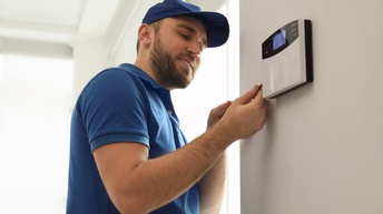 Nahaufnahme einer Person, die an einer Wand steht, an der ein kleines, rechteckiges, weißes, elektronisches Kästchen befestigt ist, auf dem drei Punkte rot leuchten. In ihrer rechten Hand hat sie ein längliches, rotes Objekt, das sie an das Kästchen hält