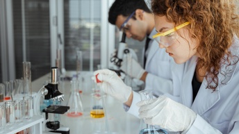 Person mit Schutzbrillen in weißem Kittel an Tisch sitzend, mit Pipette Flüssigkeit in Schälchen tropfend hält in Hand Glas mit Flüssigkeit, am Tisch Mikroskop und Röhrchen mit Flüssigkeiten, im Hintergrund weitere Person durch Mikroskop äugend