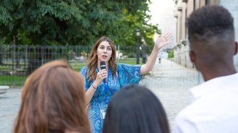 Frontale Aufnahme einer Person mit Mikrofon und Ausweisschild, die im Freien neben einem Gebäude steht und zu einer Gruppe Personen spricht. Hinter der Person ist ein Park