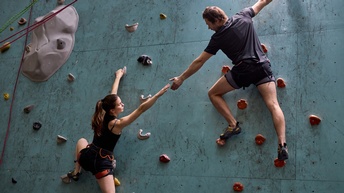 Rückenansicht zweier Personen in Sportkleidung, die mit Klettergurten in einer Kletterhalle eine Kletterwand hinaufklettern. Sie halten sich mit einem Arm fest und strecken den anderen Arm dem Gegenüber aus.