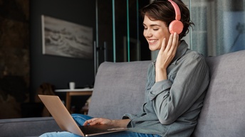 Eine Person mit braunem, kurzem Haar sowie Kopfhörern sitzt auf einem grauen Sofa. Auf ihrem Schoß ist ein aufgeklappter Laptop, auf den sie blickt
