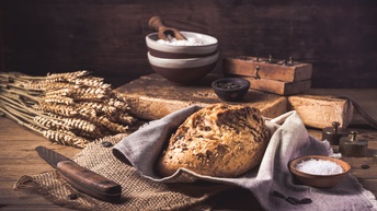 Detailansicht eines Laibes Brot auf einem hellen Tuch, umgeben von Weizenähren, einem Messer, einer kleinen Schüssel mit grobem Salz, einer Schüssel mit Mehl und einer kleinen Schüssel mit Kernen.