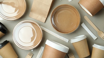 mehrere Becher mit Deckeln, Schalen mit transparenten Deckeln, Papierbeutel, einzeln verpacktes Besteck sowie Rührstäbchen. auf hellem Untergrund