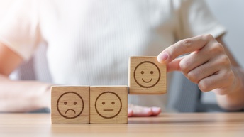 Auf einem Tisch stehen nebeneinander zwei Würfel. Auf jedem Würfel ist ein Smiley. Eine Hand hebt einen dritten Würfel in die Höhe, auf dem ein lachendes Smiley ist