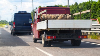 Auf einer zweispurigen, asphaltierten Straße fahren hintereinander zwei Kleintransporter. Der Hintere hat eine Ablegefläche, auf der sperrige Sachen transportiert werden