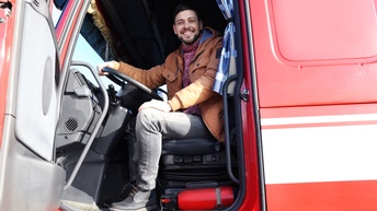 Seitliche Aufnahme einer Person mit dunklen, kurzen Haaren und Bart sowie mit dicker Jacke und Pulli, die hinter dem Lenkrad eines LKWs sitzt. Die Tür des LKWs ist geöffnet und die Person blickt direkt in die Kamera