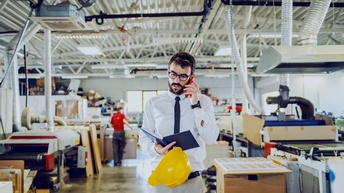 Frontale Aufnahme einer Person mit Hemd, Brille und Krawatte, die in einer Werkstatt mit Druckerpressen steht und telefoniert. Die Person hält ein aufgeklapptes Notizbuch. Im Hintergrund sind weitere Personen sowie verschiedene Drucke