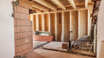 Ein Wohnraum mit Ziegelmauern, Holzwänden und einer Holzdecke. Am Boden ist Beton. In manchen Ecken liegt Schutt