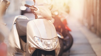 Ein weißer Motorroller von vorne. Im Mittelpunkt befinden sich die Frontverkleidung mit Scheinwerfer sowie der Lenker und der schwarze Sitz. Rechts dahinter steht ein weiterer Motorroller