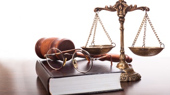 Auf einem braunen Holztisch sind Objekte platziert: Eine goldene Waage mit Verzeihrungen, ein Buch mit braunem Einband, eine Brille und ein Hammer. Der Hintergrund ist weiß.