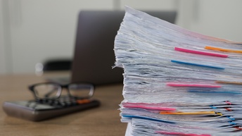 Detailansicht eines Aktenstapels, im Hintergrund verschwommen Taschenrechner mit Brille daraufliegend