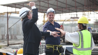 Drei Personen mit Schutzhelmen in Baustellenumgebung schlagen jubelnd die Hände aufeinander