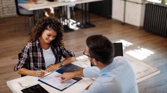 Zwei Personen sitzen sich an Tisch in Büro gegenüber, eine Person hält Stift in Händen und blickt auf vor ihr liegendes Dokument, andere Person deutet mit Hand an Stelle in Dokument