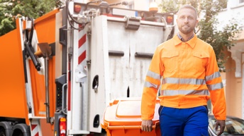 Eine Person in orange-reflektierender Jacke und blauer Hose zieht eine orange Mülltonne hinter sich her. Hinter der Person steht ein Müllauto