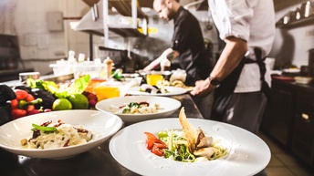 Detailansicht auf Teller in einer Gastronomieküche, ringsum kochende und Essen zubereitende Personen