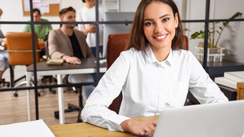 Lächelnde Person mit dunklen langen Haaren und weißer Bluse sitzt bei einem Schreibtisch und arbeitet mit einem Laptop, im Hintergrund zeigt sich eine Glastrennwand und dahinter befinden sich weitere Personen in einer Büroräumlichkeit