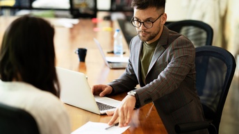 An einem Tisch sitzen zwei Personen. Die rechte Person schiebt der anderen Person einen Zettel und einen Stift hin