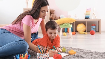 Ein Kind liegt in Bauchlage auf einem grauen Teppich. Links neben dem Kind sitzt eine erwachsene Person. Beide halten einen Stift in der rechten Hand und malen in ein offenes Heft. Vor ihnen stehen weitere Boxen mit Stiften