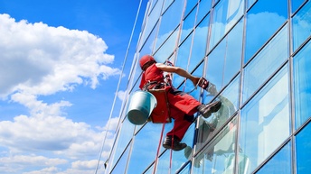 Person in roter Arbeitsausrüstung mit Helm hängt an Seilen und reinigt Fensterfassade