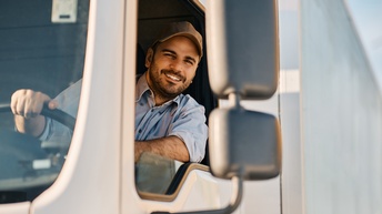 Person mit dunklen Haaren und Bart sowie brauner Kappe und blauem Hemd sitzt in einem Führerhaus eines LKWs und blickt freudig in einen Rückspiegel
