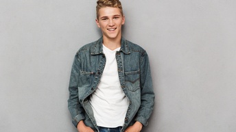 Jugendliche Person mit kurzen hellen Haaren steht freudig mit Jeansjacke und eingesteckten Händen an einer grauen Wand